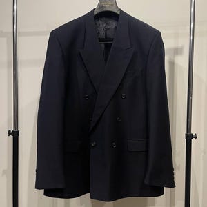May include: A dark navy double-breasted blazer on a chrome clothes rail. The jacket features a notched lapel, two rows of buttons, and two front pockets. The blazer is a solid colour and appears to be made of a woven material.