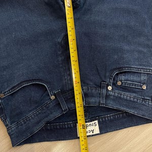May include: Close-up of dark blue denim jeans with a measuring tape showing a length of approximately 16 inches. The jeans have a classic five-pocket design and a brand label visible. The jeans are folded.