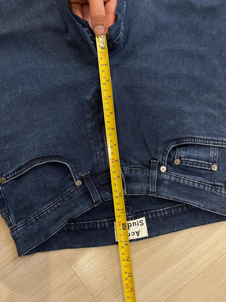 May include: Close-up of dark blue denim jeans with a measuring tape showing a length of approximately 16 inches. The jeans have a classic five-pocket design and a brand label visible. The jeans are folded.