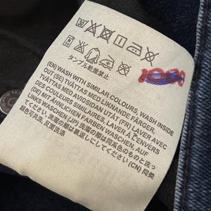 May include: Close-up of a white clothing tag with black symbols and text. The tag includes washing instructions in multiple languages, such as English, Swedish, French, German, Japanese, and Chinese. The tag is attached to dark blue denim.