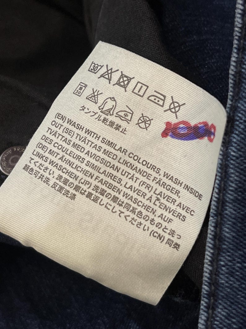 May include: Close-up of a white clothing tag with black symbols and text. The tag includes washing instructions in multiple languages, such as English, Swedish, French, German, Japanese, and Chinese. The tag is attached to dark blue denim.