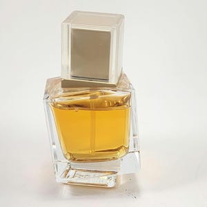 Puede incluir: Un frasco de perfume de vidrio transparente con forma cuadrada y una tapa cuadrada transparente. El perfume en su interior es de color &aacute;mbar dorado. El frasco tiene un dise&ntilde;o moderno y geom&eacute;trico.