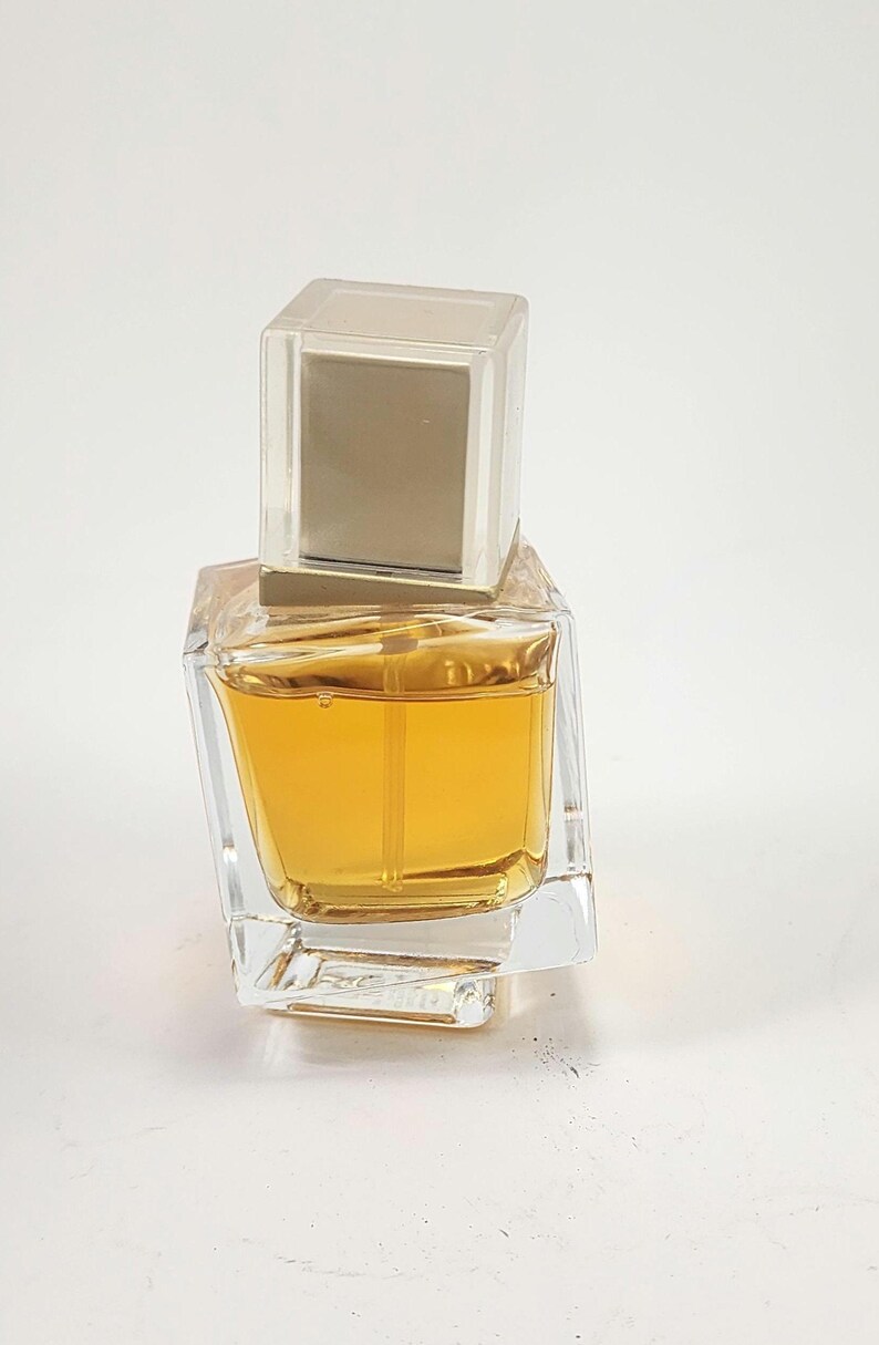 Puede incluir: Un frasco de perfume de vidrio transparente con forma cuadrada y una tapa cuadrada transparente. El perfume en su interior es de color &aacute;mbar dorado. El frasco tiene un dise&ntilde;o moderno y geom&eacute;trico.