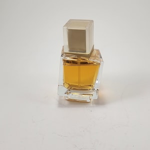 Puede incluir: Un frasco de perfume cuadrado y transparente con un l&iacute;quido de color dorado en su interior. El frasco tiene un tap&oacute;n rectangular transparente con una inserci&oacute;n dorada. El frasco de perfume est&aacute; sobre una superficie blanca.