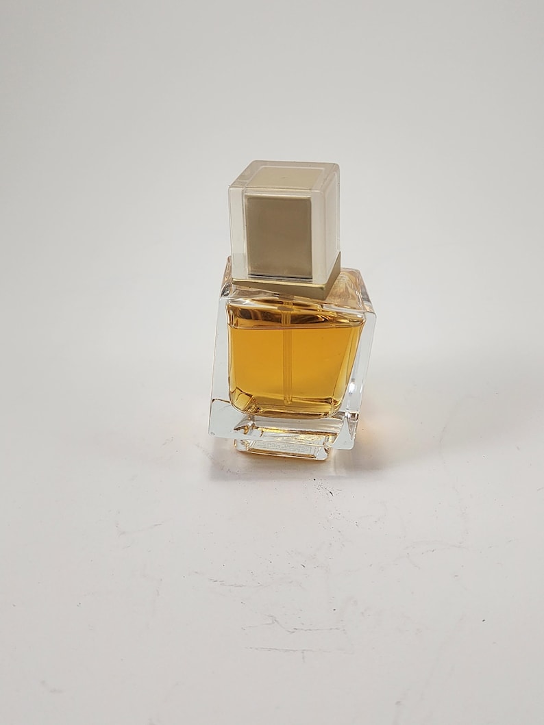 Puede incluir: Un frasco de perfume cuadrado y transparente con un l&iacute;quido de color dorado en su interior. El frasco tiene un tap&oacute;n rectangular transparente con una inserci&oacute;n dorada. El frasco de perfume est&aacute; sobre una superficie blanca.