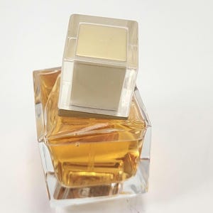 Puede incluir: Un frasco de perfume de vidrio transparente con un dise&ntilde;o geom&eacute;trico, lleno de l&iacute;quido dorado. El frasco tiene una tapa rectangular transparente con una parte superior dorada. El frasco de perfume est&aacute; sobre un fondo blanco.