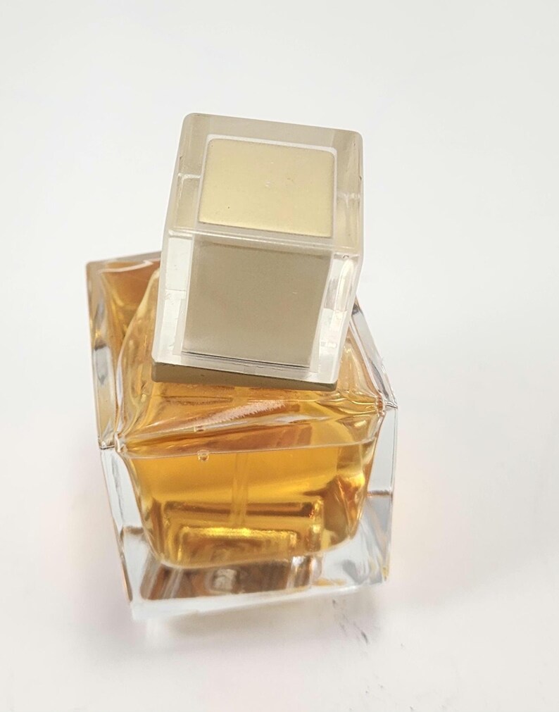 Puede incluir: Un frasco de perfume de vidrio transparente con un dise&ntilde;o geom&eacute;trico, lleno de l&iacute;quido dorado. El frasco tiene una tapa rectangular transparente con una parte superior dorada. El frasco de perfume est&aacute; sobre un fondo blanco.