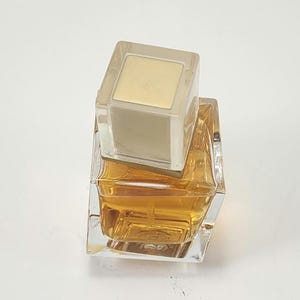 Puede incluir: Un frasco de perfume cuadrado y transparente con un l&iacute;quido dorado en su interior. El frasco tiene una tapa cuadrada y transparente con la parte superior dorada. El frasco est&aacute; sobre una superficie blanca.