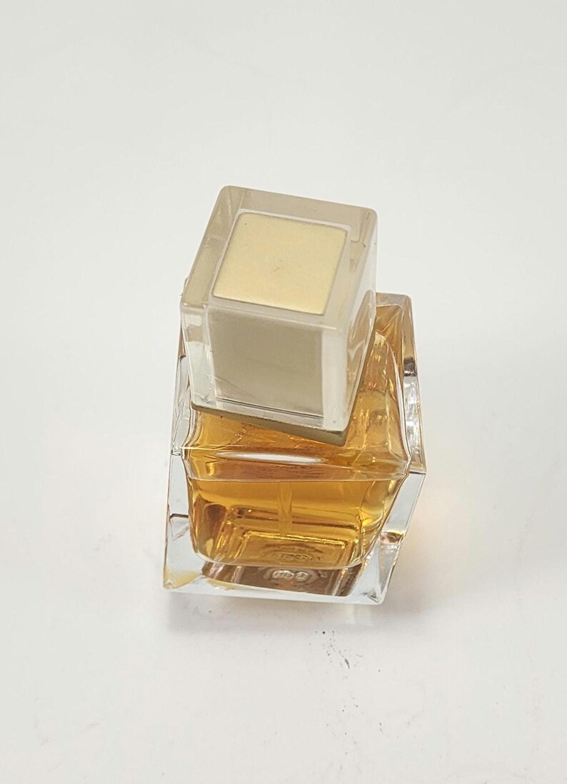 Puede incluir: Un frasco de perfume cuadrado y transparente con un l&iacute;quido dorado en su interior. El frasco tiene una tapa cuadrada y transparente con la parte superior dorada. El frasco est&aacute; sobre una superficie blanca.