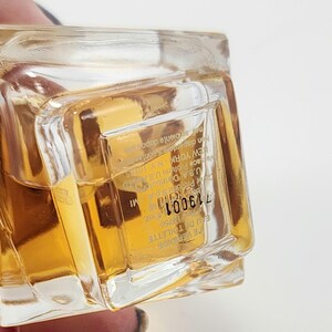 Puede incluir: Un frasco de perfume transparente y rectangular lleno de un l&iacute;quido dorado. La etiqueta incluye el texto "EAU DE TOILETTE" y el n&uacute;mero "719001". El frasco es sostenido por una persona.