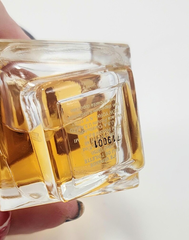 Puede incluir: Un frasco de perfume transparente y rectangular lleno de un l&iacute;quido dorado. La etiqueta incluye el texto "EAU DE TOILETTE" y el n&uacute;mero "719001". El frasco es sostenido por una persona.