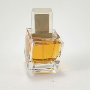Puede incluir: Un frasco de perfume de vidrio transparente con un l&iacute;quido dorado en su interior. El frasco tiene una forma angular &uacute;nica y una tapa cuadrada de color dorado. El frasco de perfume est&aacute; sobre un fondo blanco.