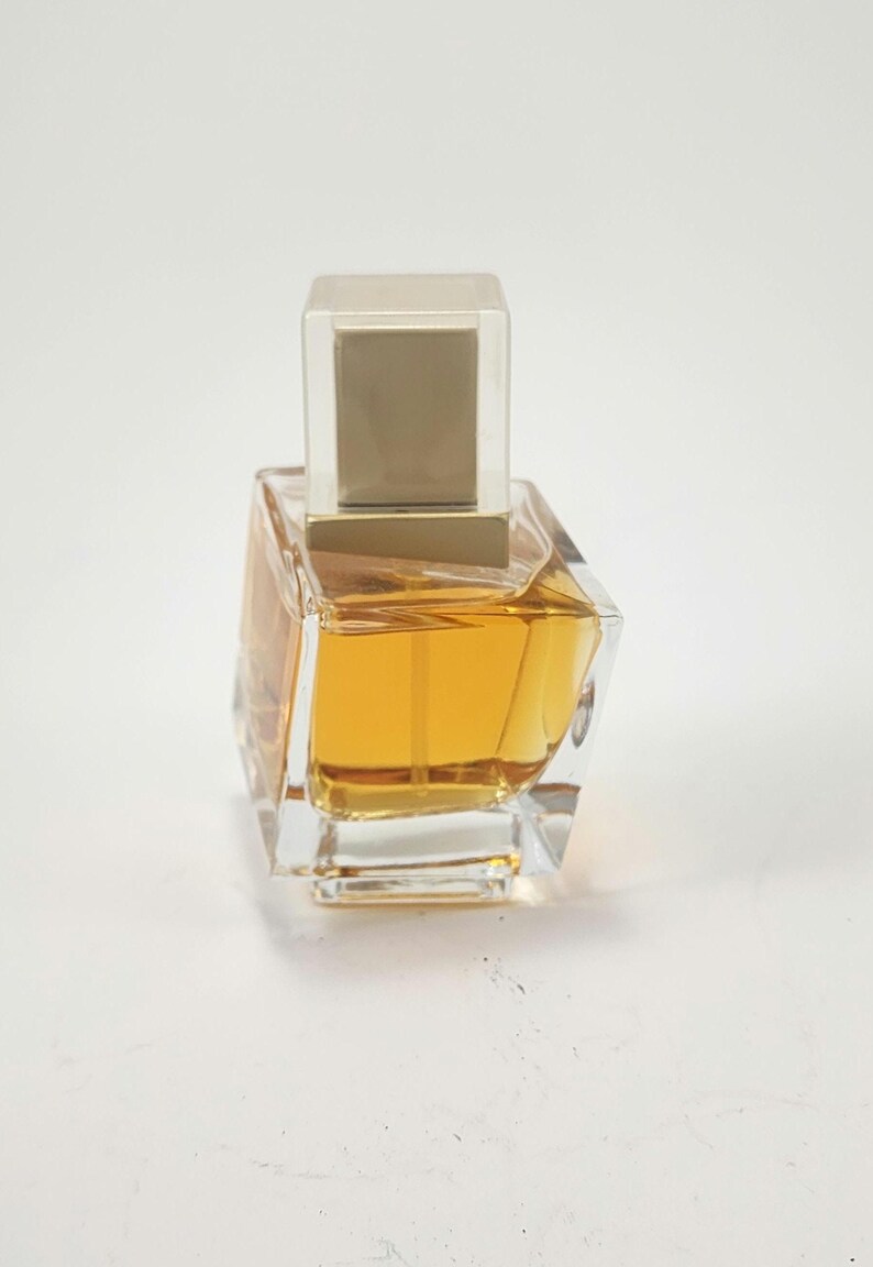 Puede incluir: Un frasco de perfume de vidrio transparente con un l&iacute;quido dorado en su interior. El frasco tiene una forma angular &uacute;nica y una tapa cuadrada de color dorado. El frasco de perfume est&aacute; sobre un fondo blanco.