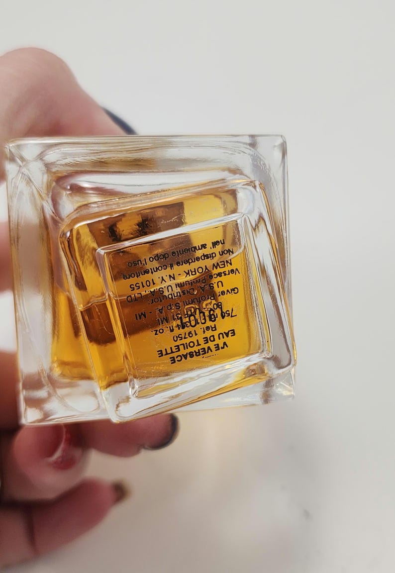 Puede incluir: Un frasco rectangular transparente de VE VERSACE Eau de Toilette. El frasco contiene un l&iacute;quido de color dorado. La etiqueta incluye el nombre de la marca, el tipo de producto y el volumen de 75 ml.