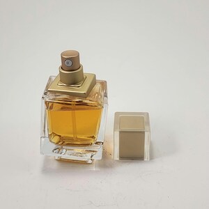 Puede incluir: Un frasco de perfume de vidrio transparente con una tapa dorada y forma cuadrada. El frasco contiene un l&iacute;quido de color dorado. La tapa est&aacute; a un lado. El frasco de perfume est&aacute; sobre una superficie blanca.
