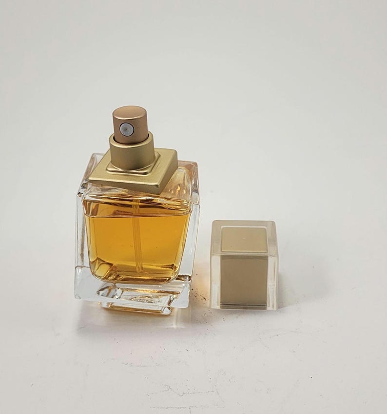 Puede incluir: Un frasco de perfume de vidrio transparente con una tapa dorada y forma cuadrada. El frasco contiene un l&iacute;quido de color dorado. La tapa est&aacute; a un lado. El frasco de perfume est&aacute; sobre una superficie blanca.