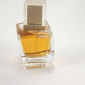 Puede incluir: Un frasco de perfume de vidrio transparente con forma cuadrada y l&iacute;quido de color dorado en su interior. El frasco tiene una tapa rectangular transparente con la parte superior dorada. El frasco est&aacute; sobre una superficie blanca.