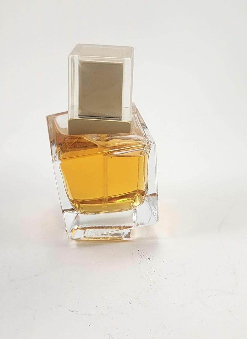 Puede incluir: Un frasco de perfume de vidrio transparente con forma cuadrada y l&iacute;quido de color dorado en su interior. El frasco tiene una tapa rectangular transparente con la parte superior dorada. El frasco est&aacute; sobre una superficie blanca.
