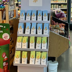 May include: A white display stand with three shelves holding rows of blue and white coffee bags. The bags have a gold logo and the text "Medium Roast Coffee". The stand is in a grocery store aisle.