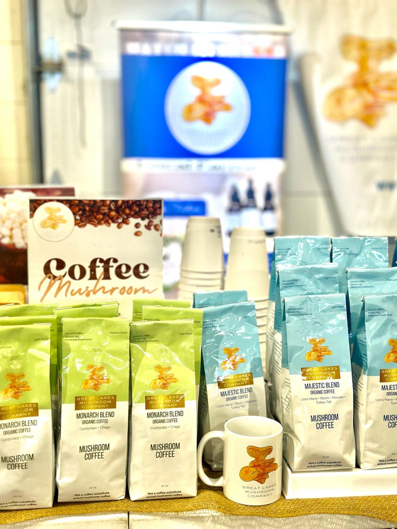 May include: A variety of coffee bags with the text "Great Lakes Mushroom Company" and "Mushroom Coffee" on the front. The bags are arranged on a table with a white mug with the same logo and a mushroom design.
