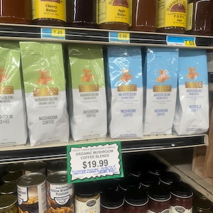 May include: A shelf display of various products, including jars of cherry butter, apple butter, and frog jam, bags of mushroom coffee, and cans of diced potatoes. A sign advertises Great Lakes Mushroom Company Organic Mushroom Coffee Blends for $19.99.