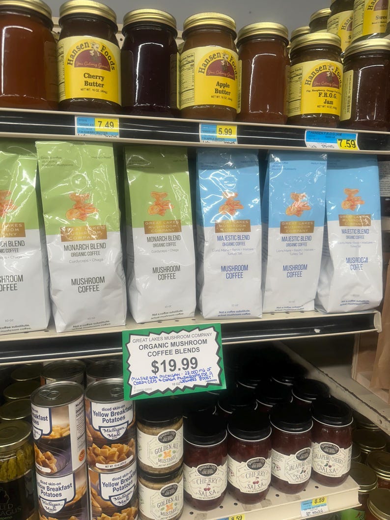 May include: A shelf display of various products, including jars of cherry butter, apple butter, and frog jam, bags of mushroom coffee, and cans of diced potatoes. A sign advertises Great Lakes Mushroom Company Organic Mushroom Coffee Blends for $19.99.