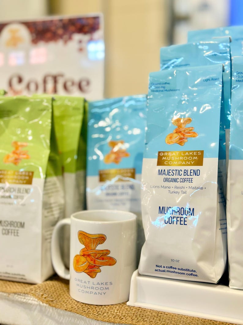 May include: A white ceramic mug with a mushroom graphic and the text "Great Lakes Mushroom Company". The mug is sitting on a table in front of several bags of coffee labeled "Majestic Blend Organic Coffee" and "Mushroom Coffee" from Great Lakes Mushroom Company.