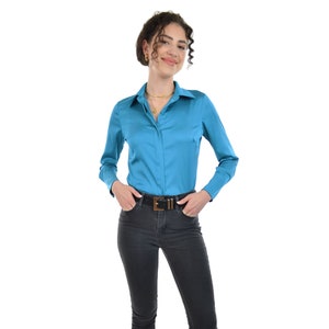 May include: A woman wearing a turquoise long-sleeve button-down shirt, black skinny jeans, and black heeled sandals. She is standing with her hands in her pockets and smiling at the camera.