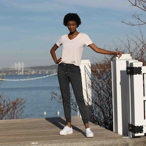 May include: A woman wearing a light pink v-neck t-shirt and dark gray sweatpants. She is standing on a wooden deck with a white fence behind her.