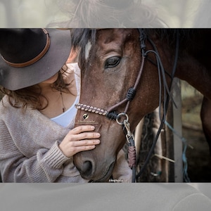 Puede incluir: Una mujer con un sombrero marrón y un suéter beige toca suavemente la nariz de un caballo marrón que lleva una cabezada de cuerda marrón y negra.