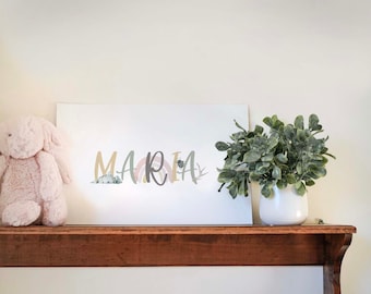 May include: A white framed print with the name "MARIA" written in colorful stylized letters with a rainbow and mountains behind the letters. The print is sitting on a wooden shelf with a potted green plant to the right.