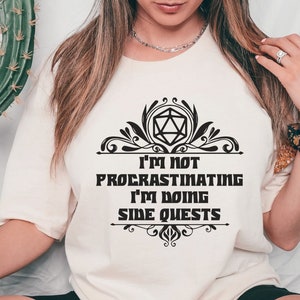 Op de afbeelding: Crèmekleurig T-shirt met de zwarte tekst "I'M NOT PROCRASTINATING I'M DOING SIDE QUESTS" en een decoratief ontwerp. Het ontwerp bevat een geometrische vorm. Dit is een casual kledingstuk.