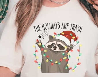 Può includere: Una maglietta bianca con una grafica di un procione che indossa un cappello di Babbo Natale e si è impigliato nelle luci di Natale. Il testo "The Holidays Are Trash" è stampato sopra il procione.