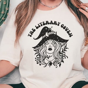 Peut inclure: Un t-shirt blanc avec un motif noir d'une sorci&egrave;re portant un chapeau orn&eacute; d'&eacute;toiles et d'un croissant de lune. Le texte "The Literary Coven" est &eacute;crit dans une police r&eacute;tro autour du motif.