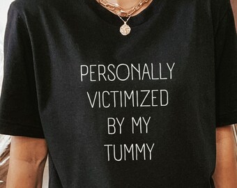 Puede incluir: Una camiseta negra con el texto "PERSONALLY VICTIMIZED BY MY TUMMY" impreso en blanco.