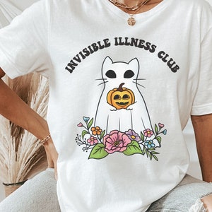 May include: White t-shirt featuring a graphic of a white cat holding a pumpkin. The text "Invisible Illness Club" is written in a retro font around the cat.