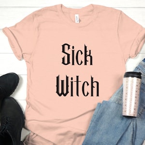 Puede incluir: Una camiseta rosa claro con el texto "Sick Witch" en fuente gótica negra. La camiseta está colocada sobre una superficie de madera blanca con un par de botas negras, unos vaqueros azules y una taza de viaje.