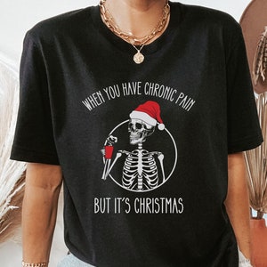 May include: Black t-shirt with a skeleton wearing a Santa hat and holding a red cup. The text on the shirt reads "When you have chronic pain but it's Christmas."