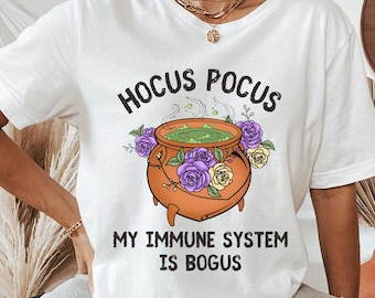 May include: White t-shirt with a graphic of a cauldron with green liquid and purple and yellow flowers. The text "Hocus Pocus My Immune System Is Bogus" is printed on the shirt.