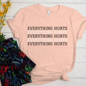 May include: A light pink t-shirt with the text "EVERYTHING HURTS" repeated five times in black and white.