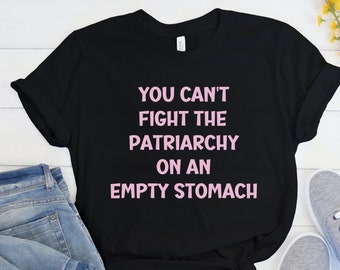 May include: Black t-shirt with the pink text "YOU CAN'T FIGHT THE PATRIARCHY ON AN EMPTY STOMACH". The shirt is shown with blue jeans and grey trainers.