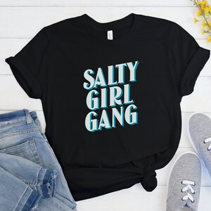 Pode incluir: Camiseta preta com o texto "Salty Girl Gang" em letras turquesa e brancas.