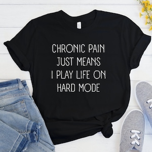 May include: Black t-shirt with white text that reads "Chronic pain just means I play life on hard mode".