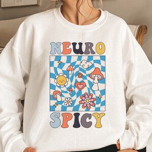 May include: White sweatshirt with a colourful graphic design featuring the text "NEURO SPICY" in a retro style. The graphic includes a chequered pattern, mushrooms, flowers, and a sun.