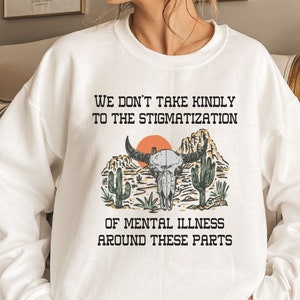以下が含まれることがあります： 「We don't take kindly to the stigmatization of mental illness around these parts.」というテキストと、頭蓋骨と交差した骨のグラフィックプリントが施された白いスウェットシャツ