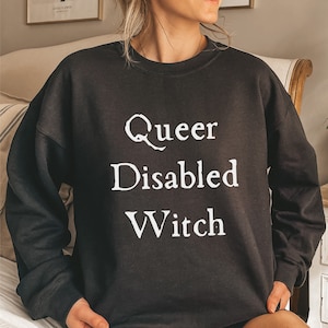 Puede incluir: Sudadera negra con texto blanco que dice "Queer Disabled Witch".