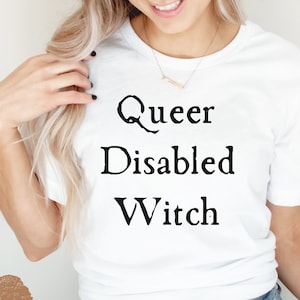 Puede incluir: Camiseta blanca con el texto "Queer Disabled Witch" impreso en negro.