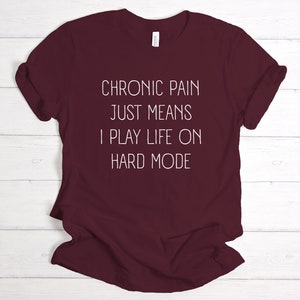 May include: Burgundy t-shirt with white text that reads "Chronic pain just means I play life on hard mode."