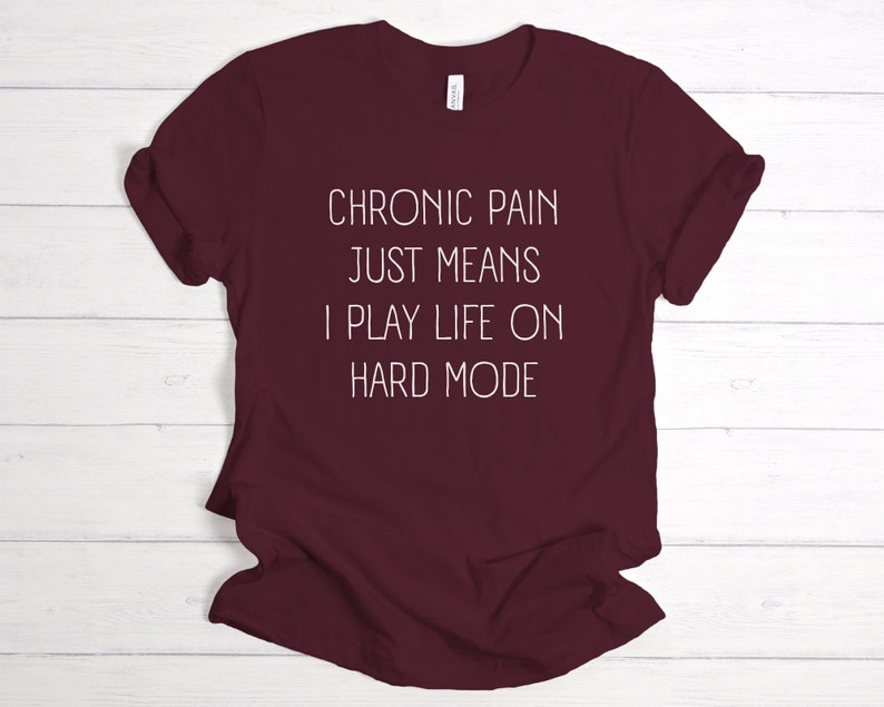 May include: Burgundy t-shirt with white text that reads "Chronic pain just means I play life on hard mode."