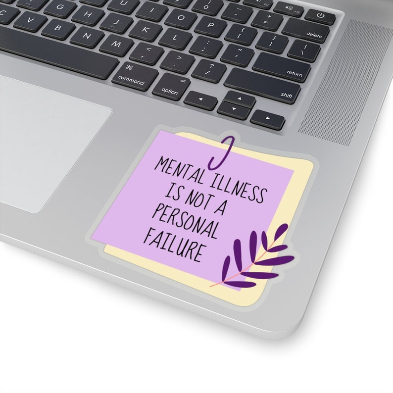 Puede incluir: Una pegatina con el texto "MENTAL ILLNESS IS NOT A PERSONAL FAILURE" en negro. La pegatina es de color lila claro con un borde amarillo y un dise&ntilde;o de hojas moradas. Un clip plateado sujeta la pegatina.